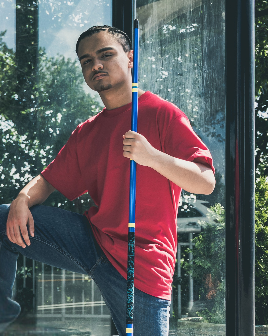 Man in a red shirt holding a blue pole against a glass window with reflections of trees.