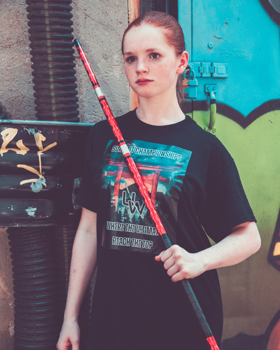 Person wearing a black t-shirt with graphic design, holding a red and black object against a graffiti-covered wall.